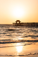 Naples, Florida yellow and orange sunset in gulf of Mexico with sun setting by Pier wooden jetty, with horizon and dark silhouette ocean waves, sunlight reflection