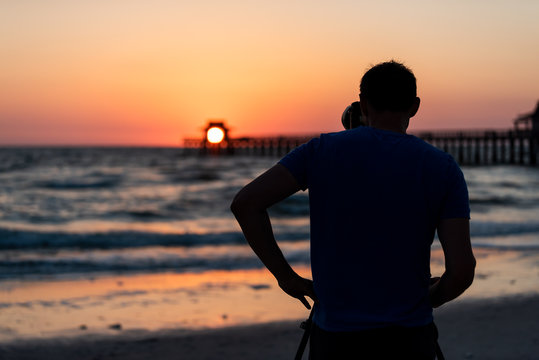 Naples, Florida red and orange sunset in gulf of Mexico with sun setting inside Pier bokeh, back of young man photographer videographer taking pictures filming landscape