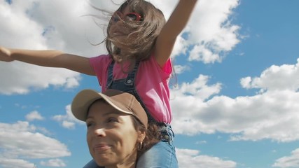 Circling the child. A child plays with mother in nature. Fly with mom. A happy little girl sits on her mother's shoulders with her hands up.