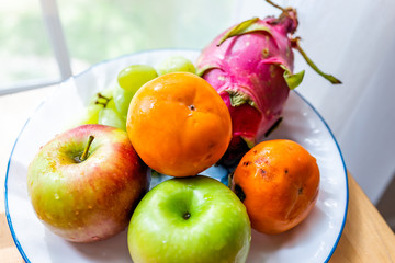Closeup of whole orange, green, red dragon dragonfruit fruit fruits, apples, persimmons kaki, on plate, wooden table by window, autumn season