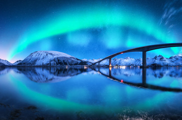 Fototapeta na wymiar Bridge and aurora borealis over snowy mountains. Lofoten islands, Norway. Amazing northern lights and reflection in water. Winter landscape with starry sky, polar lights, road, sea, city illumination