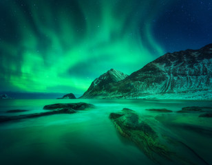 Fototapeta na wymiar Aurora borealis above snowy mountain and sandy beach with stones. Northern lights in Lofoten islands, Norway. Starry sky with polar lights. Night winter landscape with aurora, sea with blurred water