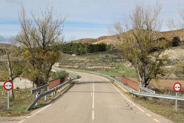 A paved two ways street going into the Aragonese countryside hills, with bare trees and green...