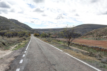 A paved two ways street going into the Aragonese countryside hills, with bare trees and green...