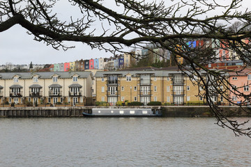 Tree branches and a moored flatboat in the Avon river wharf, in front of beige and colored terraced houses, during winter, in Bristol, United Kingdom