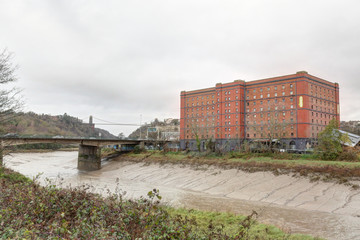 Obraz premium The Avon river with mud after a storm, next to a red brick abandoned industrial building and a bridge in a cloudy winter in Bristol, United Kingdom