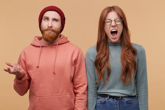 indoor shot of young male with confused facial expression, looks above and his girlfriend standing near him, open mouth widely and scream.