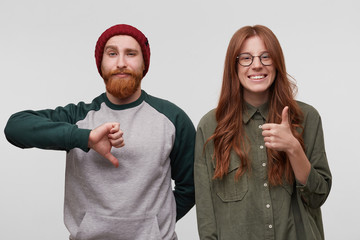 Charming redhead young woman smiles and thumbs up, because she like something. Her boyfriend near her feels unhappy and shows thumbs down gesture. Isolated over white wall © timtimphoto