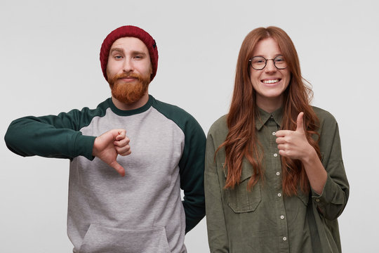 Charming redhead young woman smiles and thumbs up, because she like something. Her boyfriend near her feels unhappy and shows thumbs down gesture. Isolated over white wall