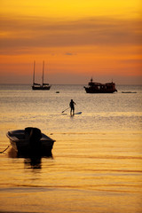 man standing on sup ,stand up paddle board sailing in koh tao harbor against beautiful sunset sky