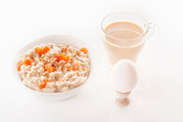 Fototapeta na wymiar Oatmeal with pumpkin and nuts in a plate, a glass of tea, a boiled egg and a jug with milk on a white background. Close-up.