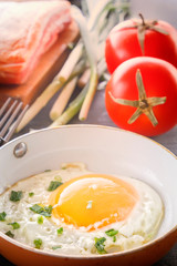 Fried egg in a pan, sliced bread, bacon, tomatoes and green onions cooked for breakfast on a wooden gray table. Close-up