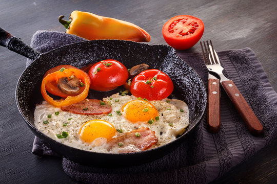 Fried eggs with bacon and tomatoes on an old cast iron pan and cutlery on a gray table. Close-up