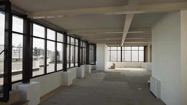 Camera shows Large empty hall with big black framed windows, grey floor and white walls and ceiling without any furniture and people in daylight.