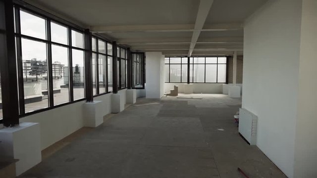 Camera shows Large empty room with big black framed windows, grey floor and white walls and ceiling without any furniture and people in daylight.