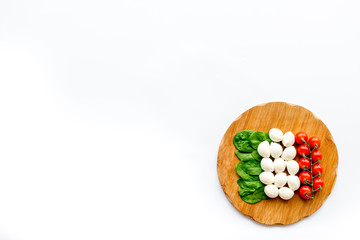 Italian flag made of mozzarella cheese, cherry tomatoes, green basil on wooden cutting board on white background top view copy space