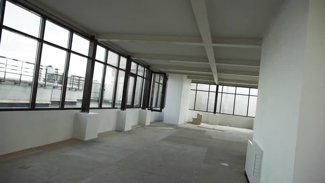 Camera shows large empty room in apartment with big black framed windows, grey floor and white walls and ceiling without any furniture and people in daylight.
