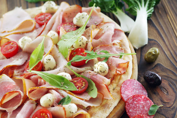 Pizza on a wooden table close-up with prosciutto ham, mozzarella, cherry tomatoes and various ingredients.
