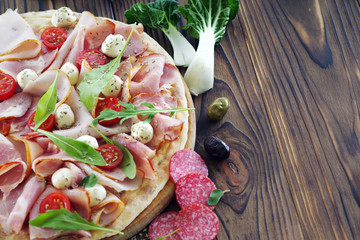 Pizza on a wooden table, with prosciutto ham, mozzarella and various ingredients.
