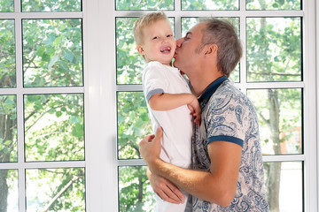 Fototapeta premium Adult man sitting on window sill carrying happy little boy and kissing him in cheek