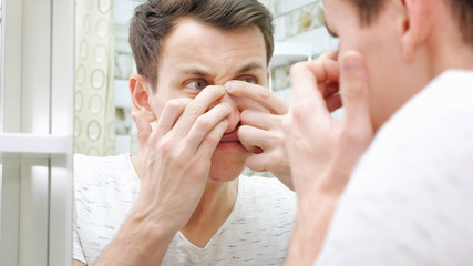 close up of young man standing close to a mirror in the bathroom and squeezing a pimple on his face.