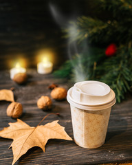cup of coffee in paper cup on wooden table with blurred pine tree leaves and candles in background