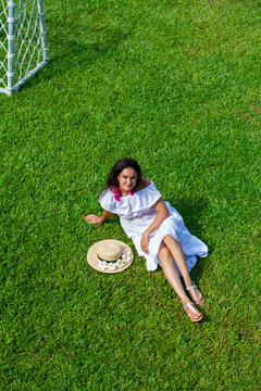 Brunette Girl In A Straw Hat Sits On A Green Lawn With Frangipani Flowers Next To The Football Goal