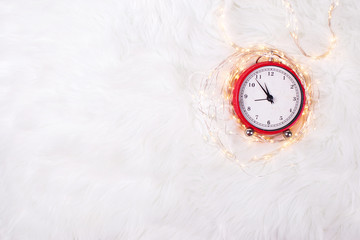 Red  alarm clock - symbol of  New Year and fairy lights  on white fur background.