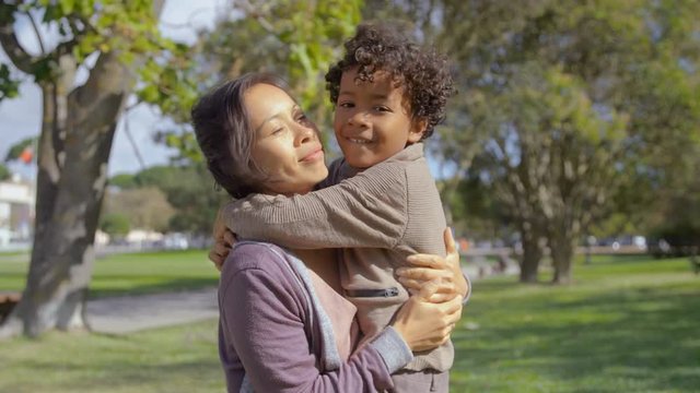 Curly African American son hugging and kissing mother in slow motion. Cheerful Asian mom embracing little girl in summer park. Parenthood concept