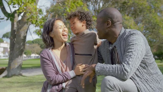 Cheerful Multicultural parents hugging and kissing son on green lawn with sunlight. Portrait of happy Interracial family with kid in park in slow motion. Family concept
