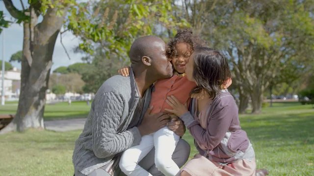 Mixed race daughter hugging and kissing parents in park with sunlight. Portrait of happy Interracial family with kid in park in slow motion. Loving concept