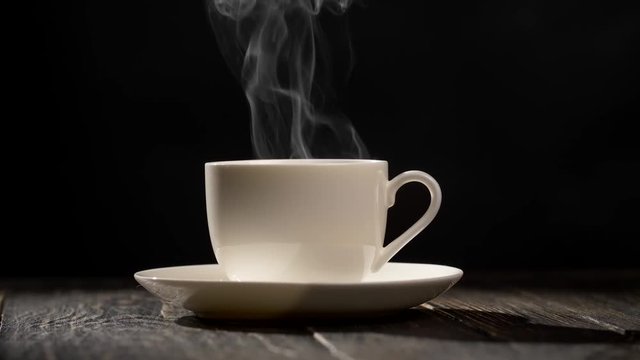 Cup of hot coffee. Steam coming out of a coffee cup standing on a white saucer on a wooden table. Close-up shot with a black background, UHD