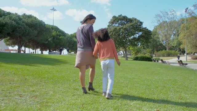 Cutly daughter walking and holding hands with mother in green park. Rear view of mom and girl walking in park in slow motion. Motherhood concept