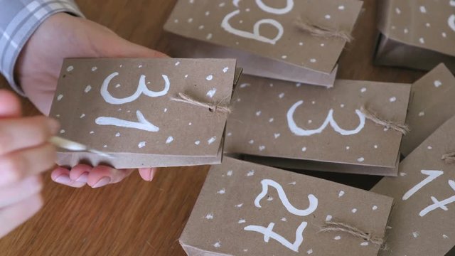 Woman makes christmas advent calendar for kids. Writes a number on the bag by brush and white paint. Close-up hands.