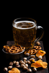 Glass of lager beer with snack on vintage wooden board on black background. Pistachios and pretzel with potato crisps.