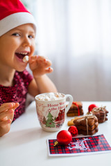 Little girl in Santa hat eating marshmallow from mug with cacao and cookies, Merry Cristmas
