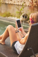 Girl using cellphone while lying on a swimming pool deck lounge bed.