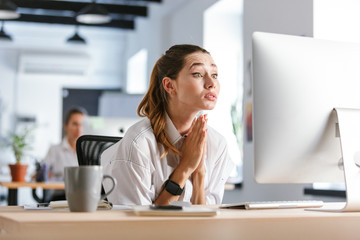 Worried young businesswoman sitting at her workplace at the office, looking at computer, begging