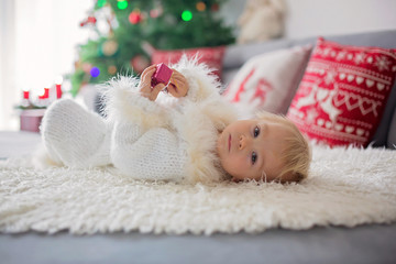 Curious little toddler boy, dressed in handknitted white overall, lying on the couch playing with gifts