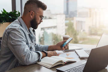 Hipster man sits in cafe, uses smartphone, works on two laptops. Businessman reads an information message in phone.