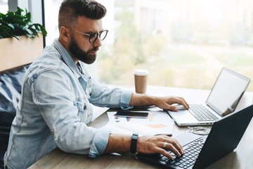Side view. Hipster bearded businessman sits in cafe, works on two laptops. Freelancer work remotely. Online education.