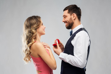 love, couple, proposal and people concept - happy man giving diamond engagement ring in little red box to woman over grey background