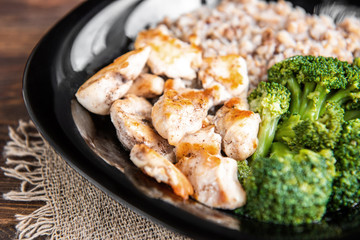 Roasted chicken breast with broccoli and buckwheat porridge on black plate on wooden background.