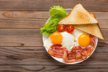 White plate of fried eggs with bacon, toasts and vegetables on wooden table, top view. Copy space