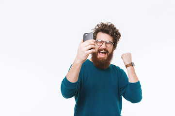 Naklejka premium Amazed and cheerful bearded man looking at smartphone and celebrating success over white background