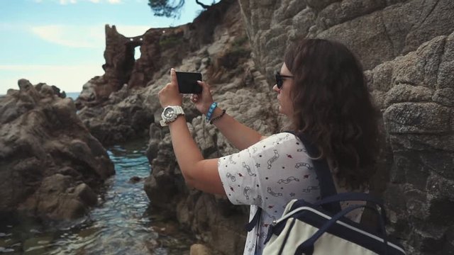 View from behind of a brunette female tourist with backpack taking photos of rocks cliffs and sea. Beautiful nature view, visiting touristic places.