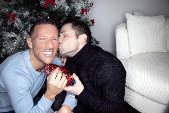 Gay Male Couple Exchanging Christmas Gift In Front Of Tree With A Kiss