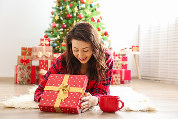 Obraz na płótnie Canvas Young beautiful brunette woman in plaid checkered pajamas in decorated bedroom interior with gift box and Chrictmas tree on background. People on Christmas morning concept. Close up, copy space.