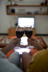 Crop shot of couple relaxing at home and toasting with wineglasses on sofa against burning TV