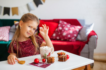 Little girl in red dress eating Christmas cookies with cacao in cup, red Chirstmas decorations around